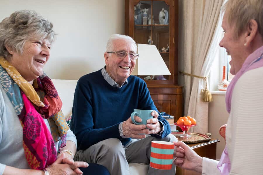 Three seniors sit in the living room of the senior villa home, laughing and talking and drinking coffee