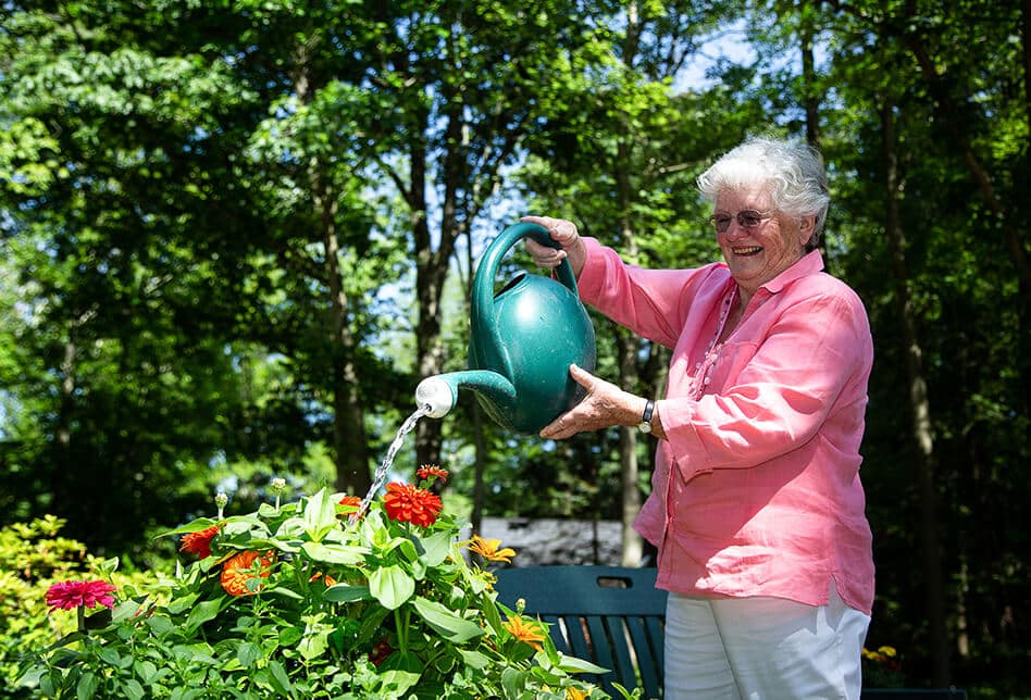 An elderly woman watering flowers outside while smiling.