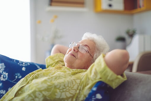 An elderly woman puts her hands behind her head and leans back on the couch and closes her eyes