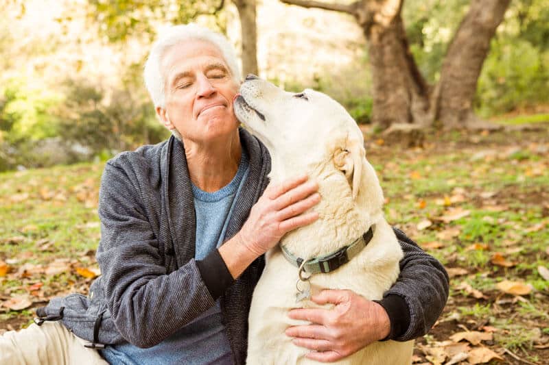 A senior man smiling while he hugs his dog from the side and the dog licks his cheek.
