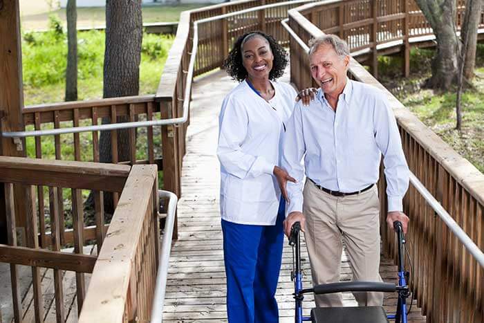 A senior man with a walker goes on a walk on a boardwalk with the assistance of a health care professional