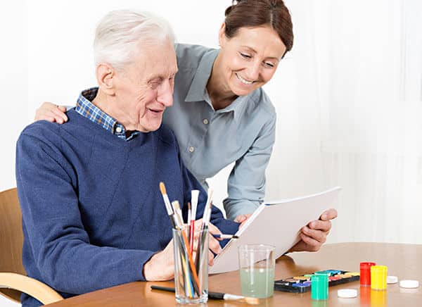 An elderly man admiring his painting while an instructor smiles over his shoulder.